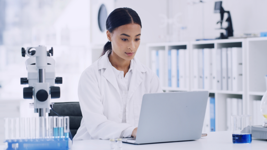 Young woman scientist in white lab coat working on laptop computer at laboratory bench surrounded by test tubes with blue liquid solutions and microscope equipment in bright modern research facility, demonstrating digital healthcare technology integration in medical research