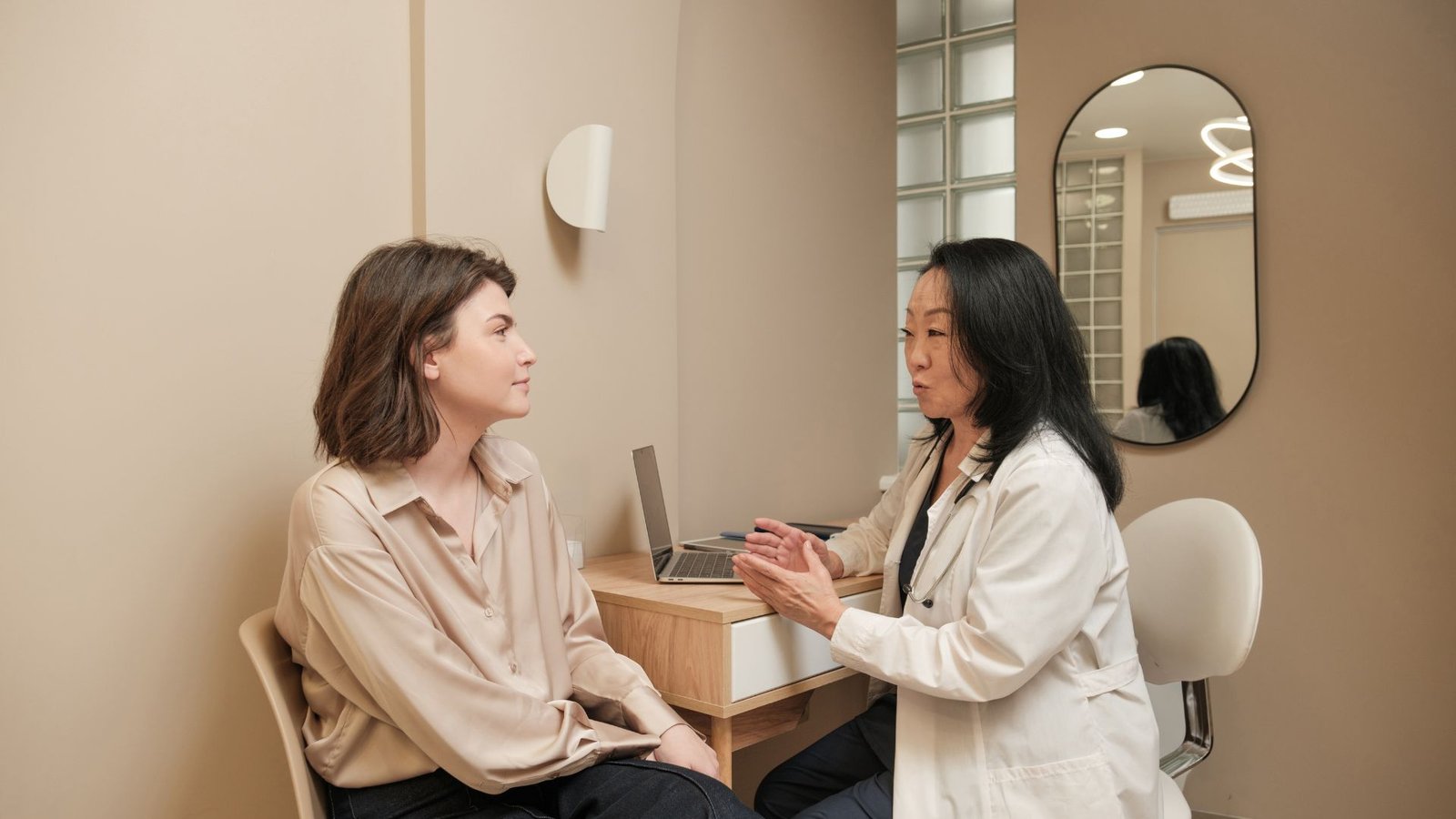 Female medical professional in white coat gesturing during consultation with attentive patient in contemporary minimalist examination room with arch mirror and soft neutral tones