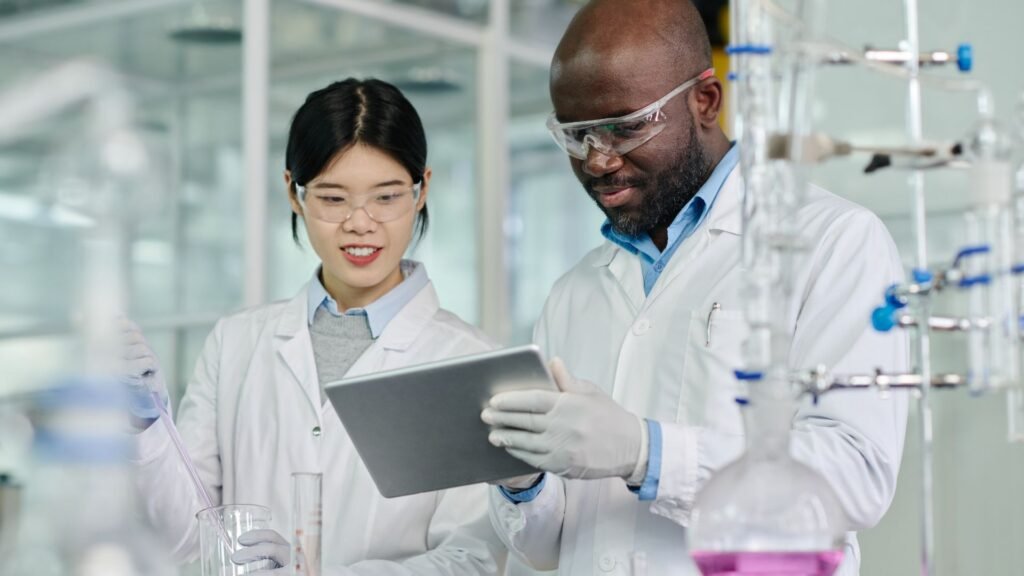 female scientist and a male scientist in white lab coats and safety goggles review information on a digital tablet while standing at a laboratory workstation. Laboratory glassware and equipment are visible around them.