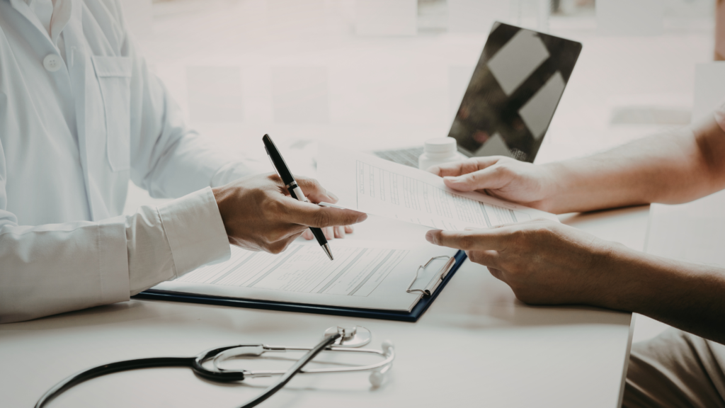 Healthcare provider in white coat pointing to medical documents on clipboard while patient holds tablet device and reviews paperwork during clinical consultation, stethoscope visible on desk