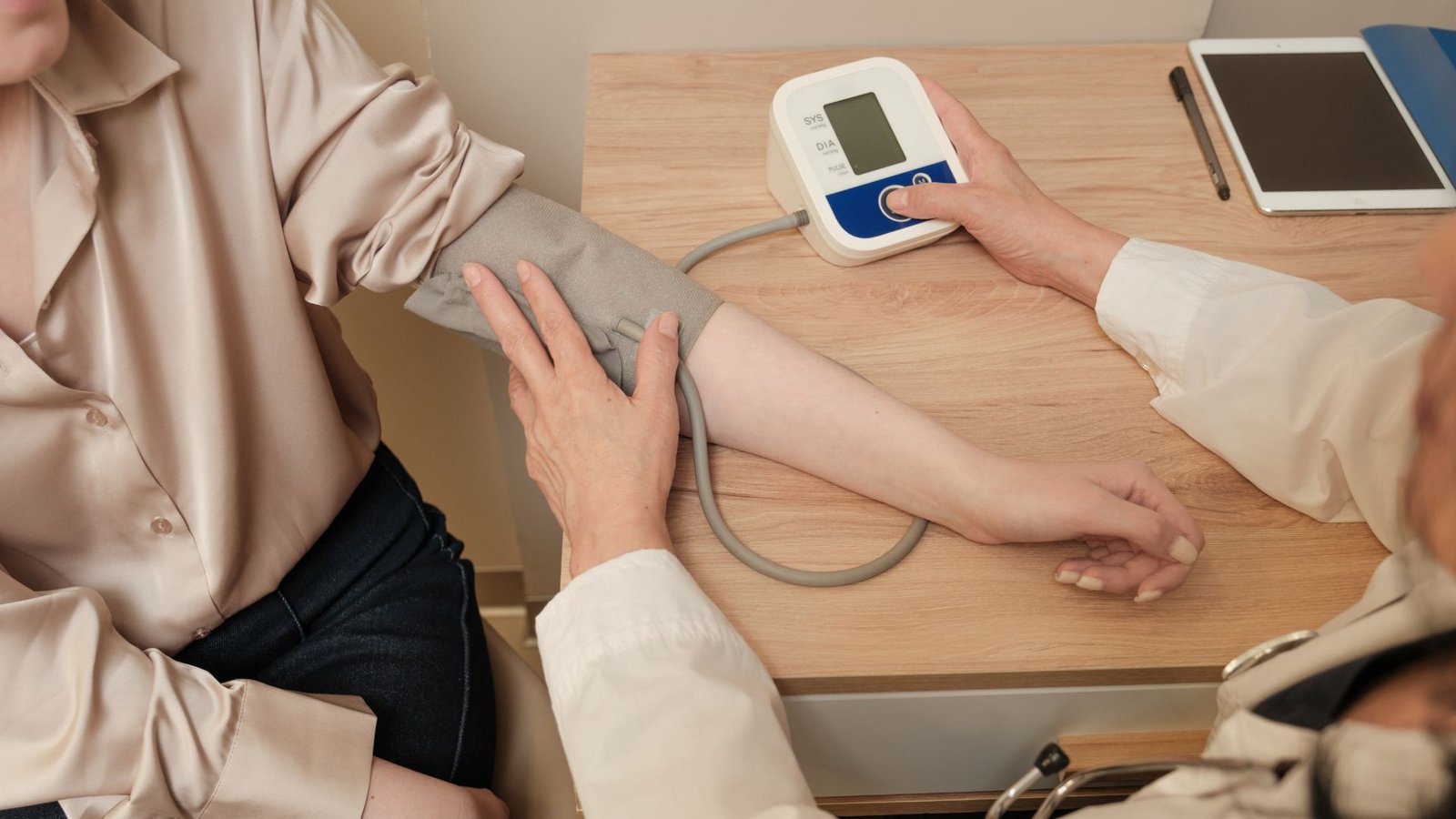 Person in beige blouse using digital blood pressure monitor with arm cuff on wooden desk alongside tablet and notebook, demonstrating home health monitoring and chronic condition management