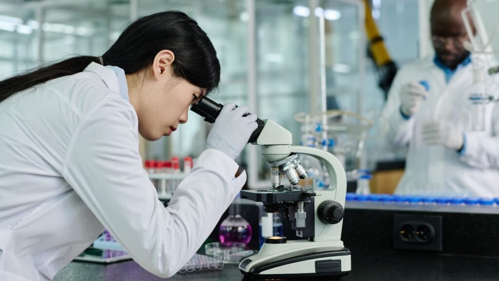 woman in a white lab coat and protective gloves focuses intently while looking through a professional microscope in a modern laboratory. Laboratory equipment and test tubes in various colors are visible in the background.