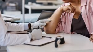 Medical professional in white coat and gloves preparing to administer injection into upper arm of patient wearing pink shirt at clinical desk with medical equipment and paperwork, demonstrating preventive healthcare and immunization services