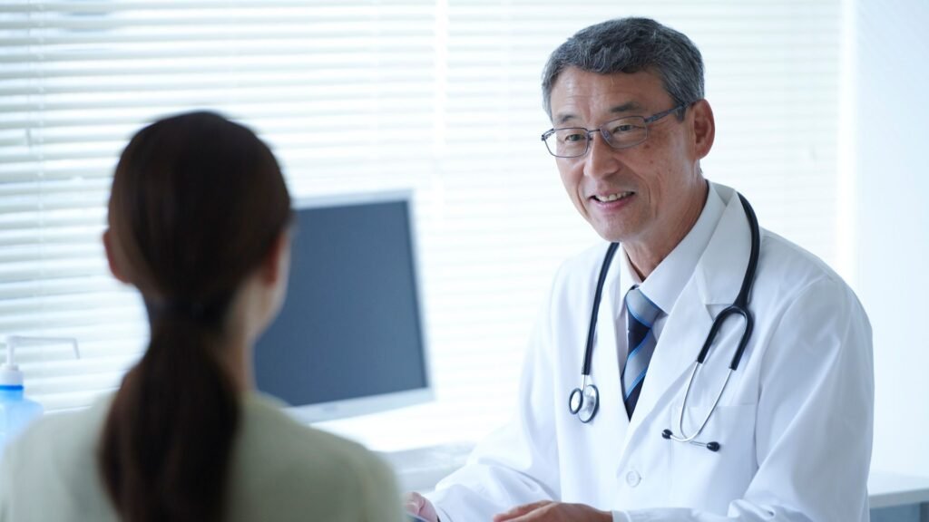 Smiling male doctor with glasses and stethoscope in white coat meeting with female patient near computer in naturally lit medical office, conveying trust and professional patient care