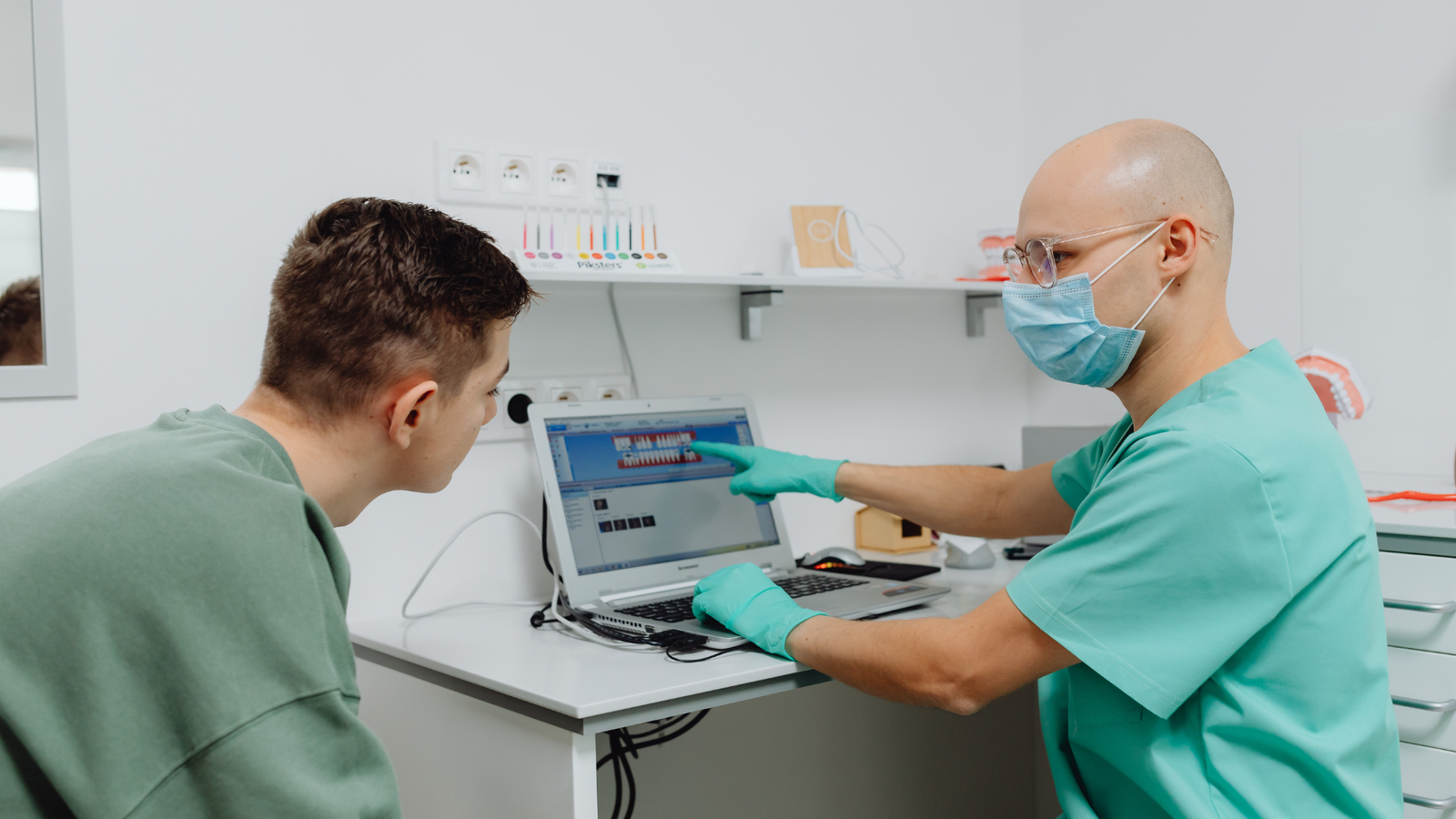 Dentist wearing teal scrubs, surgical mask, and glasses pointing to dental imaging on laptop screen while consulting with male patient in green shirt in modern dental clinic, showing use of digital technology in patient education and treatment planning