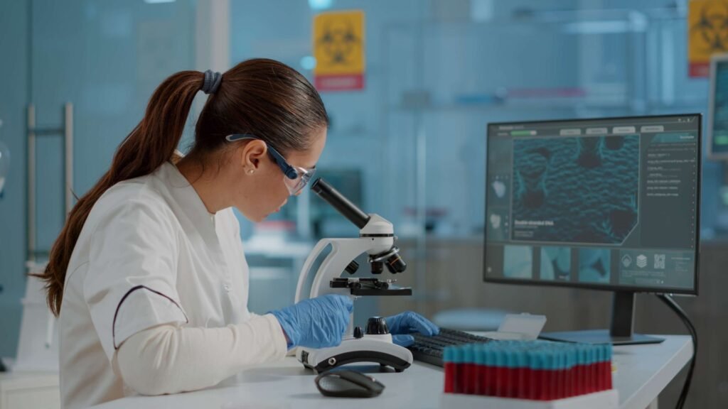 A female laboratory technician wearing protective eyewear and a white lab coat examines samples under a microscope, with blue-gloved hands carefully handling the equipment. Test tubes and a computer monitor displaying data are visible in the background.