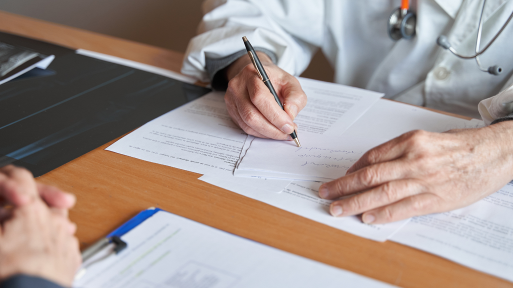 Close-up of physician's hands in white coat signing medical documents at a desk with stethoscope visible, demonstrating healthcare administrative responsibilities