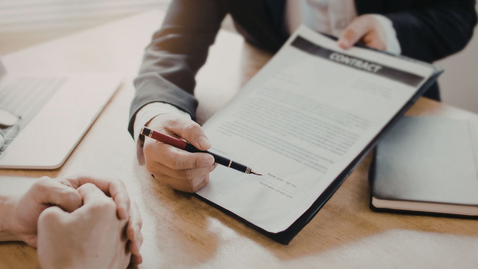 Business professional in grey suit holding pen and pointing to document on clipboard during consultation meeting at wooden desk with tablet and papers, representing contract review or medical informed consent process