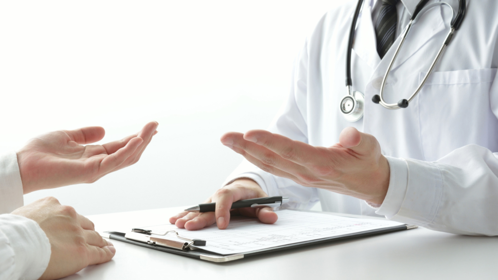 Doctor in white coat with stethoscope using hand gestures to explain medical information to patient across desk with clipboard and pen in bright clinical setting, demonstrating clear communication and shared decision-making in healthcare