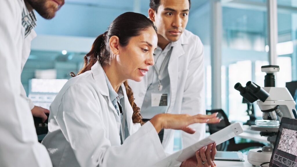 Three scientists in white lab coats collaborate in a modern laboratory setting, with a female researcher in the foreground reviewing documents while two male colleagues observe. A microscope is visible on the workbench beside them.