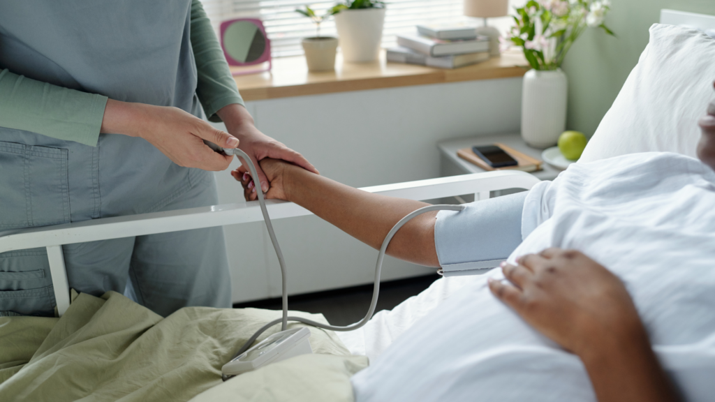 Healthcare professional in grey scrubs using a blood pressure monitor on a pregnant patient's arm in a comfortable home bedroom setting with natural light and houseplants