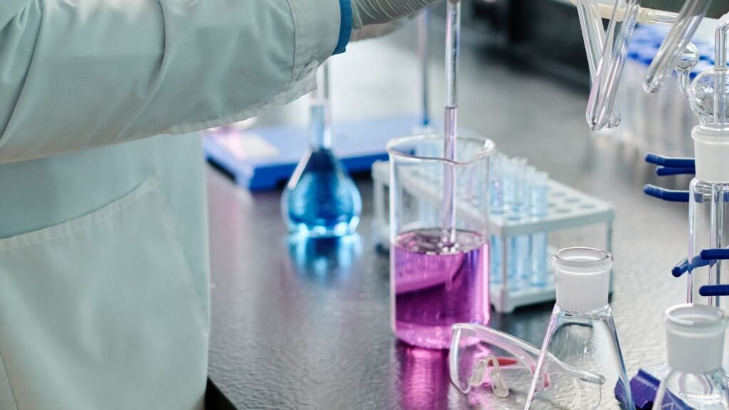 Researcher in light blue protective lab coat working with colorful chemical solutions including pink/purple liquid in beakers and blue flask alongside clear glassware in professional laboratory setting, demonstrating scientific testing and analysis