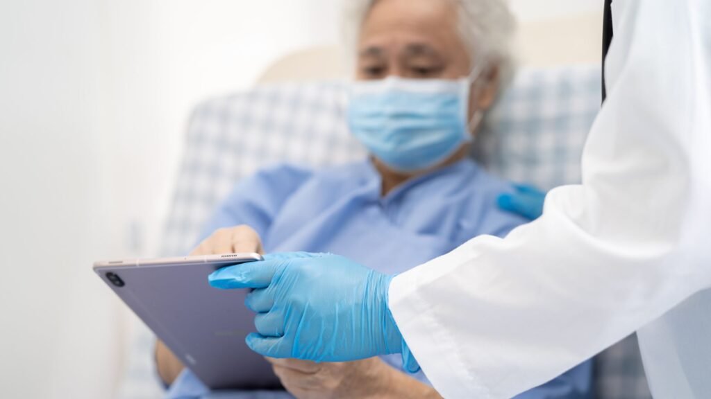 Healthcare provider in white coat and blue gloves holding tablet device while consulting with elderly patient wearing surgical mask in clinical setting, showing modern digital healthcare delivery