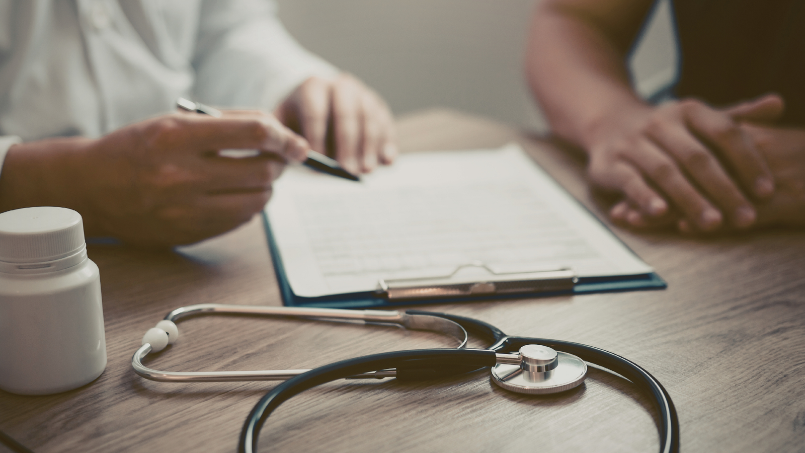 Close-up of medical consultation with healthcare professional writing notes on clipboard at wooden desk while patient gestures during discussion, stethoscope and prescription medication bottle visible in foreground, representing thorough patient assessment and documentation