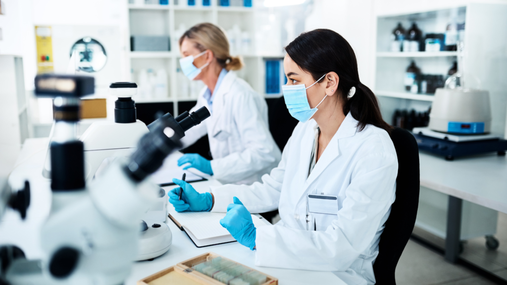 Two female lab technicians in white coats, blue surgical masks, and protective gloves conducting microscopic research at laboratory workstation with multiple microscopes, sample slides, and notebooks in bright modern clinical laboratory with organized shelving in background