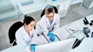 Blog Posts 5 Overhead view of two female scientists in white lab coats and blue protective gloves working together at a clean white desk. The senior researcher wearing glasses points at a tablet screen while consulting with her younger colleague. A microscope, laptop, clipboard, and other laboratory equipment are visible on the workspace in a modern research facility.