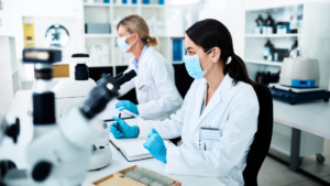 Two female lab technicians in white coats, blue surgical masks, and protective gloves conducting microscopic research at laboratory workstation with multiple microscopes, sample slides, and notebooks in bright modern clinical laboratory with organized shelving in background