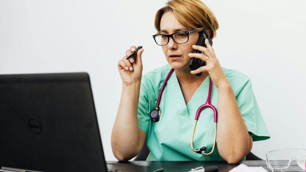 Nurse in mint green scrubs with pink stethoscope wearing glasses and holding pen while speaking on phone at desk with laptop, demonstrating telehealth or patient communication responsibilities
