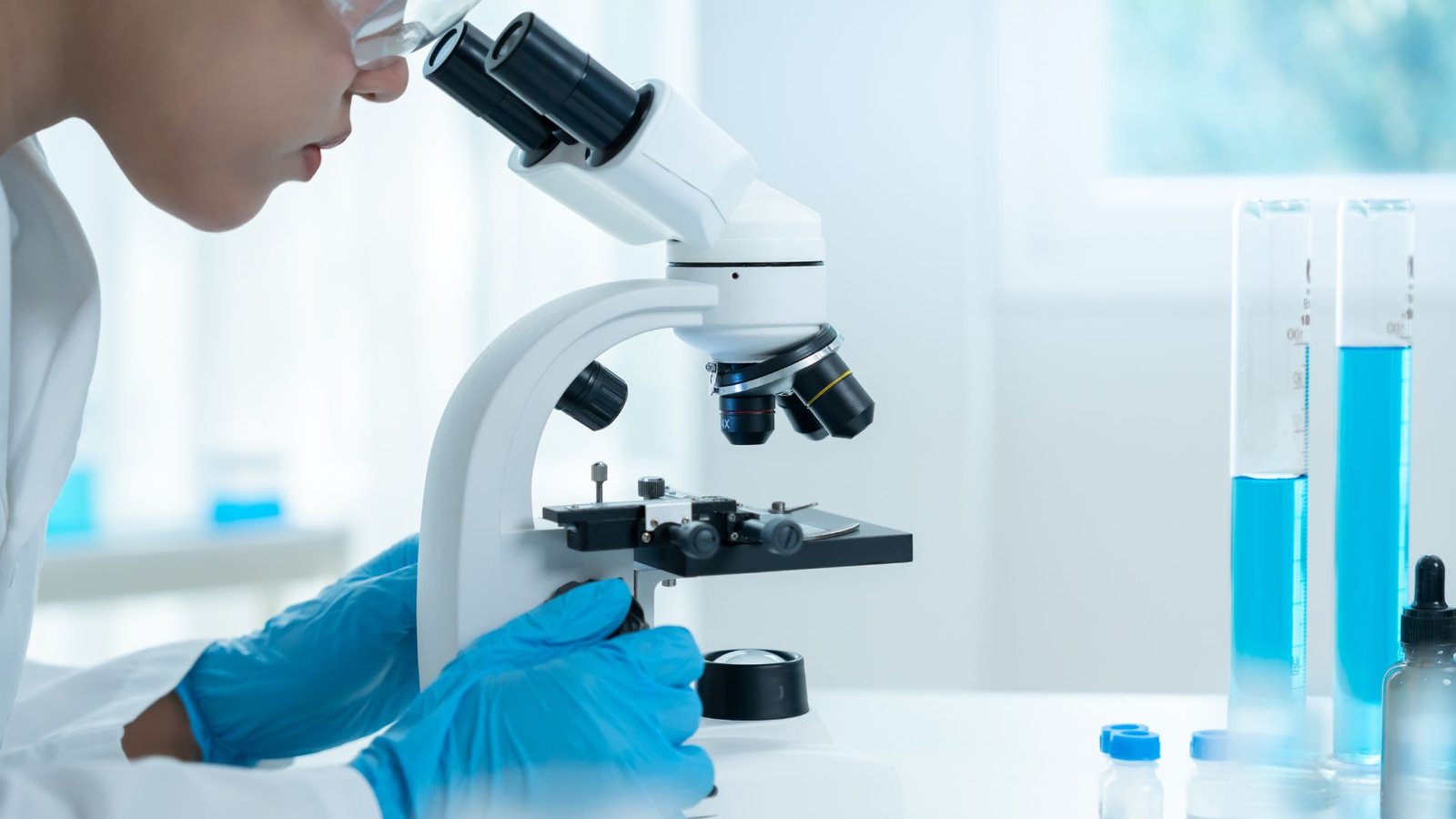 A scientist wearing protective blue gloves and a white lab coat examines a sample under a white microscope. Test tubes containing blue liquid and laboratory equipment are arranged on the clean workstation in a bright research facility.