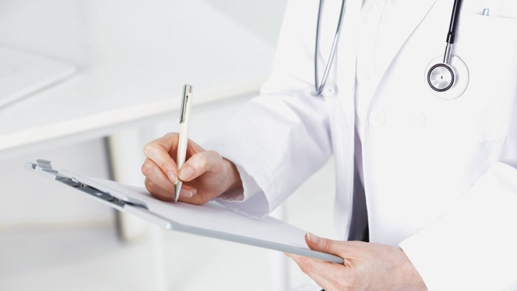 Close-up of healthcare professional in white coat with stethoscope writing on clipboard with pen in clean clinical environment, representing medical documentation and patient record-keeping