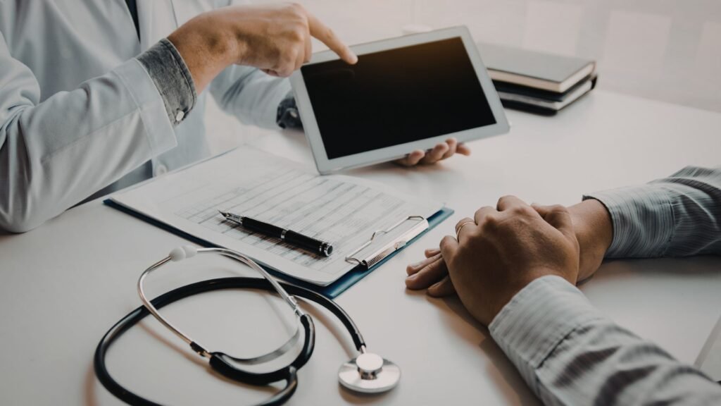 A doctor in a white coat holds and points to a digital tablet while consulting with a patient across a desk. A stethoscope, clipboard with medical forms, and other clinical materials are visible on the white consultation table.