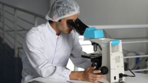Blog Posts 5 A male laboratory technician wearing a white lab coat and protective hair net conducts microscopic analysis using a blue and white microscope in a sterile laboratory environment. Documents are visible on the workspace beside him.