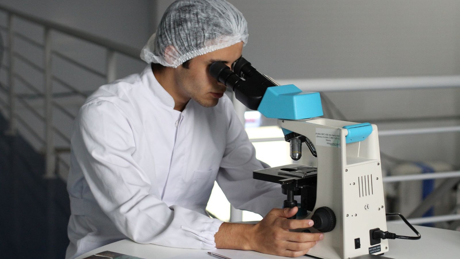 A male laboratory technician wearing a white lab coat and protective hair net conducts microscopic analysis using a blue and white microscope in a sterile laboratory environment. Documents are visible on the workspace beside him.
