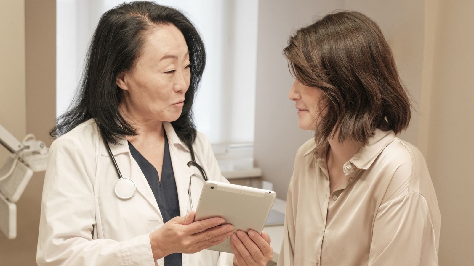 Healthcare provider in white coat with stethoscope holding tablet device while consulting with female patient in cream blouse in bright modern medical examination room, demonstrating digital health record review and patient-centered communication in contemporary clinical practice