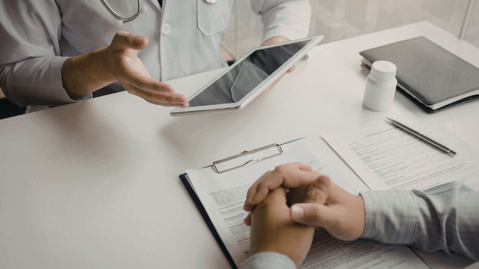 Medical professional in white coat with stethoscope showing information on digital tablet to patient across clean white desk with medical documents, prescription bottle, notebook and pen, representing modern patient education and shared decision-making in healthcare consultation