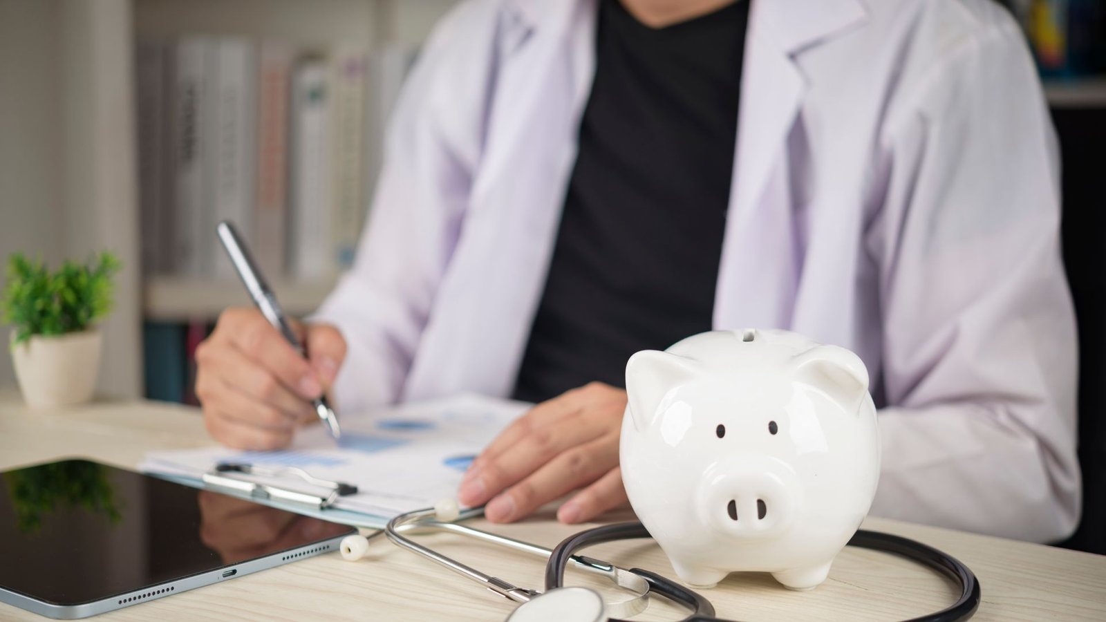 Healthcare worker in light purple blazer and black top writing notes on financial documents at desk with white piggy bank, stethoscope, tablet, and small potted plant, representing healthcare cost management, medical billing, insurance planning, and financial wellness in clinical practice
