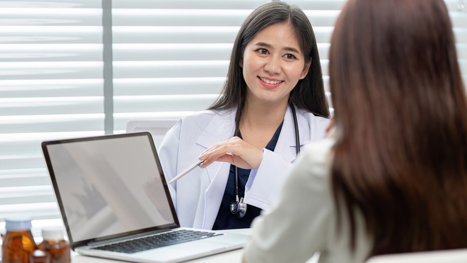 Smiling female physician in white coat with stethoscope gesturing toward laptop screen while consulting with patient in bright modern medical office with window blinds, prescription bottles visible on desk, demonstrating digital healthcare communication and patient engagement