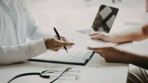 Healthcare provider in white coat writing on medical forms while patient holds X-ray film and paperwork during clinical consultation, stethoscope visible on desk, demonstrating comprehensive patient care including diagnostic assessment and treatment planning