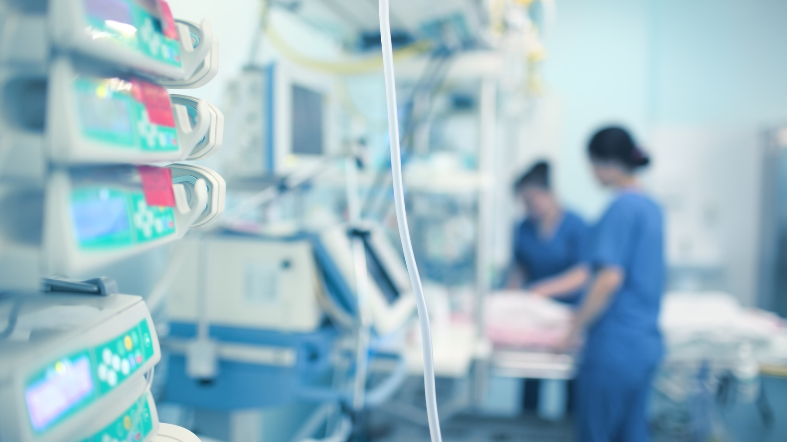 Blurred hospital intensive care unit scene showing medical monitoring equipment with digital displays in the foreground and healthcare professionals in blue scrubs attending to a patient in the background
