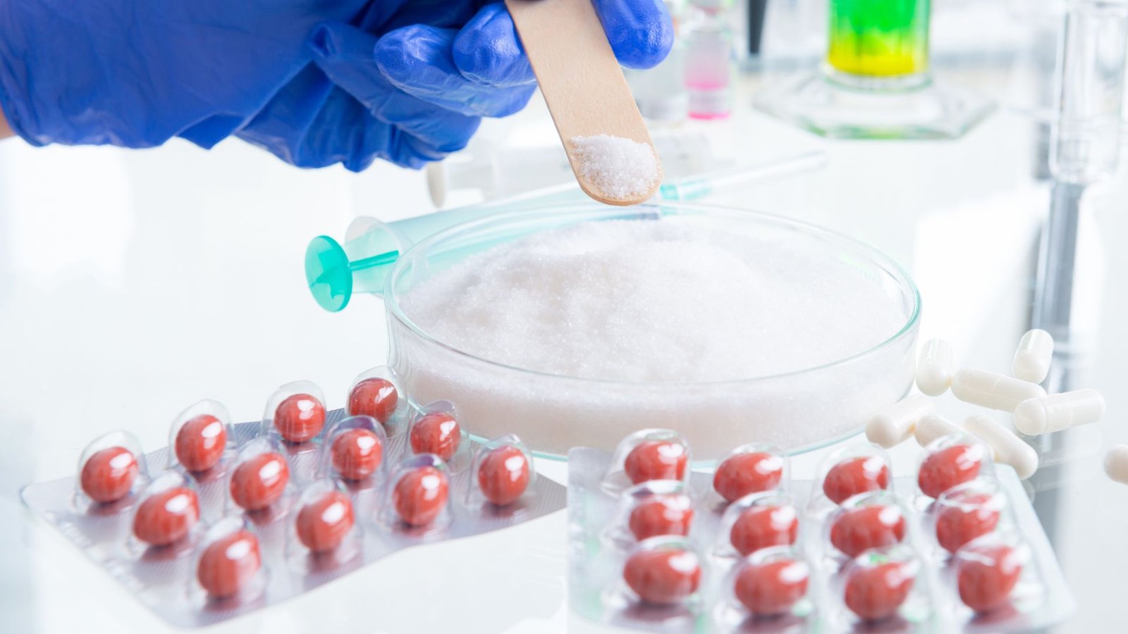 A person in blue gloves is seen adding a powder-like substance to a petri dish in a laboratory setting, with capsules and syringes visible around. This suggests the preparation or testing of pharmaceutical products in a controlled environment.