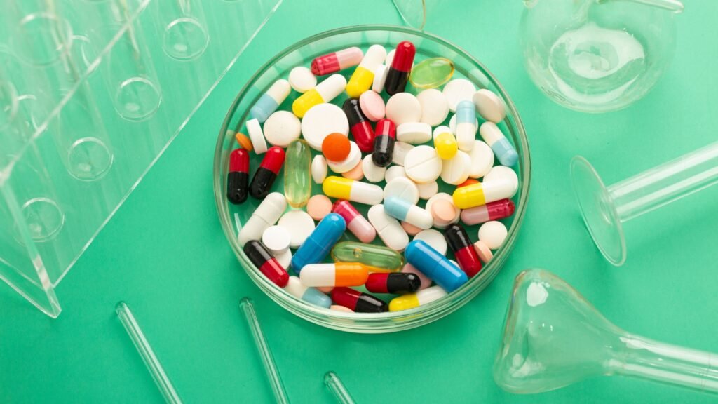 A wide variety of colorful pills are placed in a petri dish, surrounded by laboratory glassware.