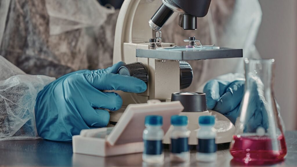 A close-up view of a scientist in protective gloves adjusting the focus of a microscope, with small laboratory vials and a beaker of red liquid in the foreground. The environment is clearly a research laboratory.