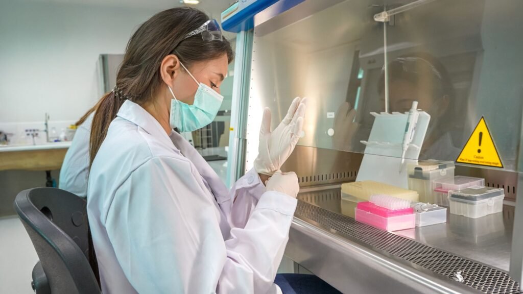 A scientist in a lab coat and face mask carefully working with a pipette inside a biosafety cabinet. The workspace is organized with colorful lab containers and equipment.