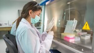 A scientist in a lab coat and face mask carefully working with a pipette inside a biosafety cabinet. The workspace is organized with colorful lab containers and equipment.