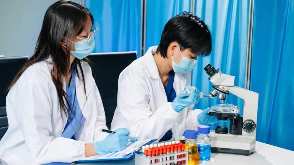 A female and male scientist in white lab coats and blue gloves working together in a laboratory, one looking into a microscope and the other taking notes. Vials and liquid samples are visible on the table.