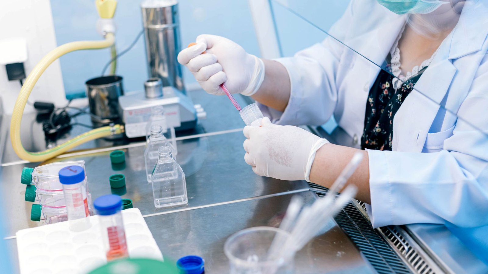 A close-up of a scientist wearing protective gloves and a mask, mixing liquid samples in a laboratory setting. The workspace is filled with laboratory equipment and plastic containers.
