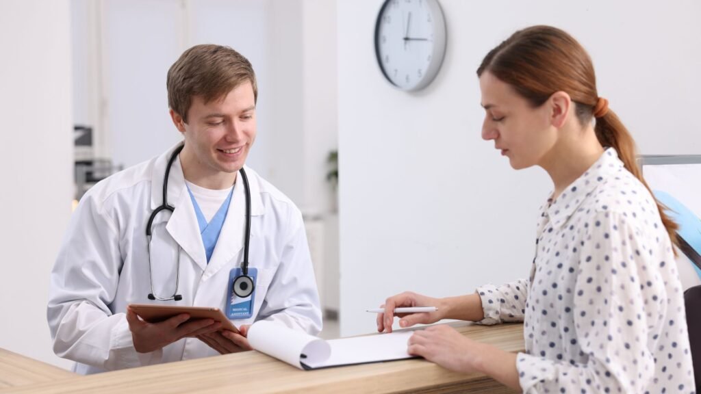 A medical assistant smiles as she assists a patient, handing them paperwork at the reception area of a healthcare facility.
