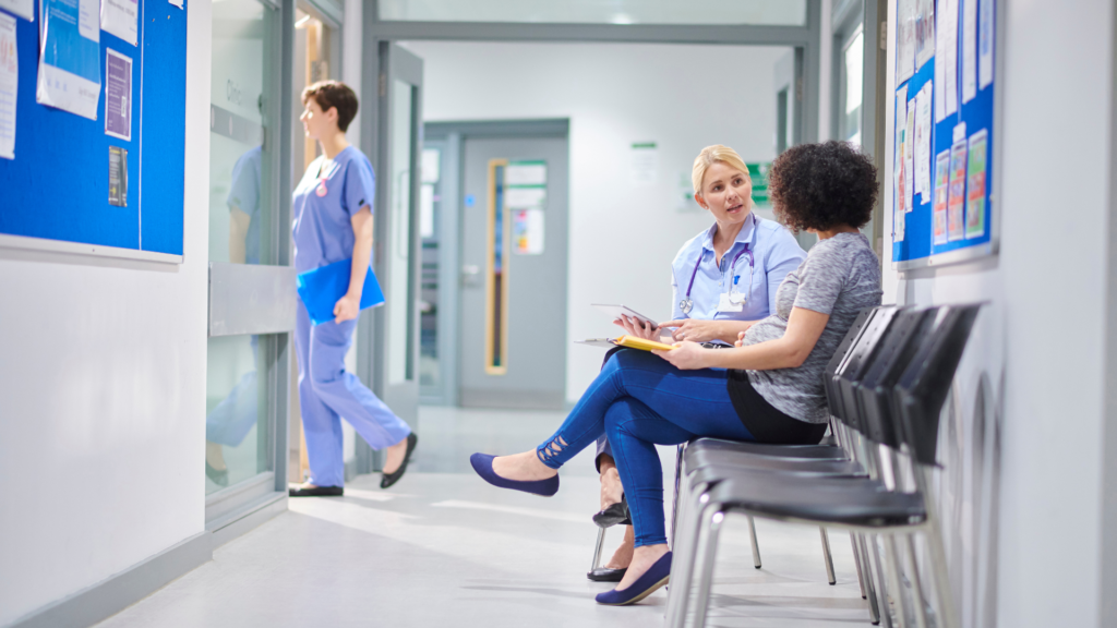 A healthcare professional and a pregnant woman sit in a hospital corridor, having a conversation while a healthcare worker walks by.