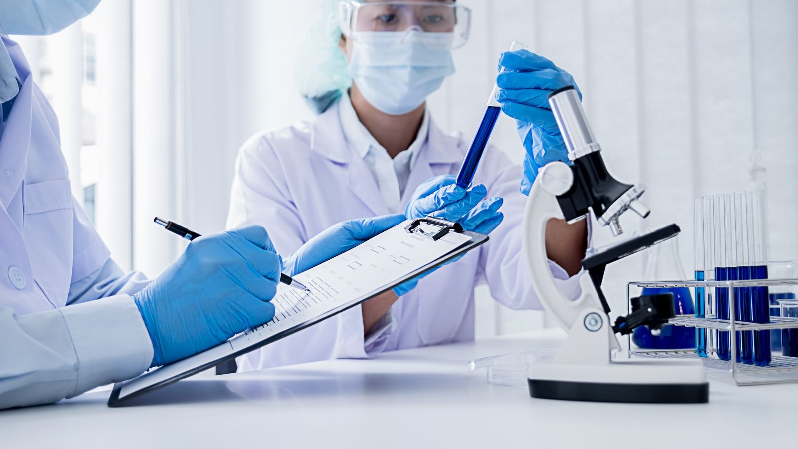 Two scientists in protective gear work together in a laboratory, one recording data while the other handles a test tube.