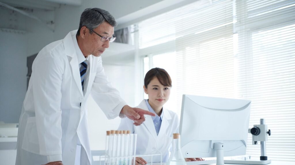An elderly doctor and a younger female doctor are reviewing data on a computer screen in a bright, modern medical office.